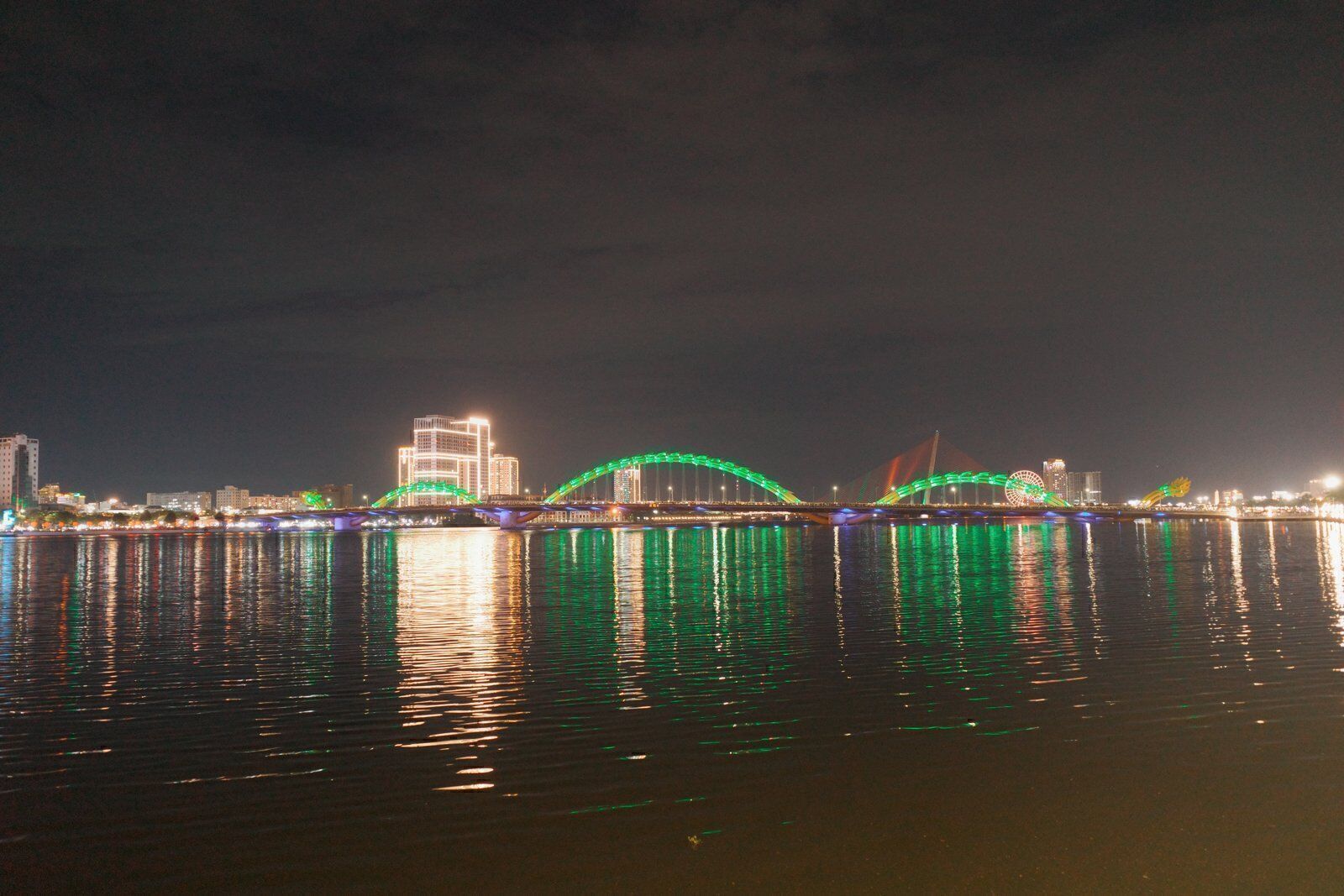 Dragon Bridge lit up in green at night across the Han River in Da Nang with the city skyline reflected in the water
