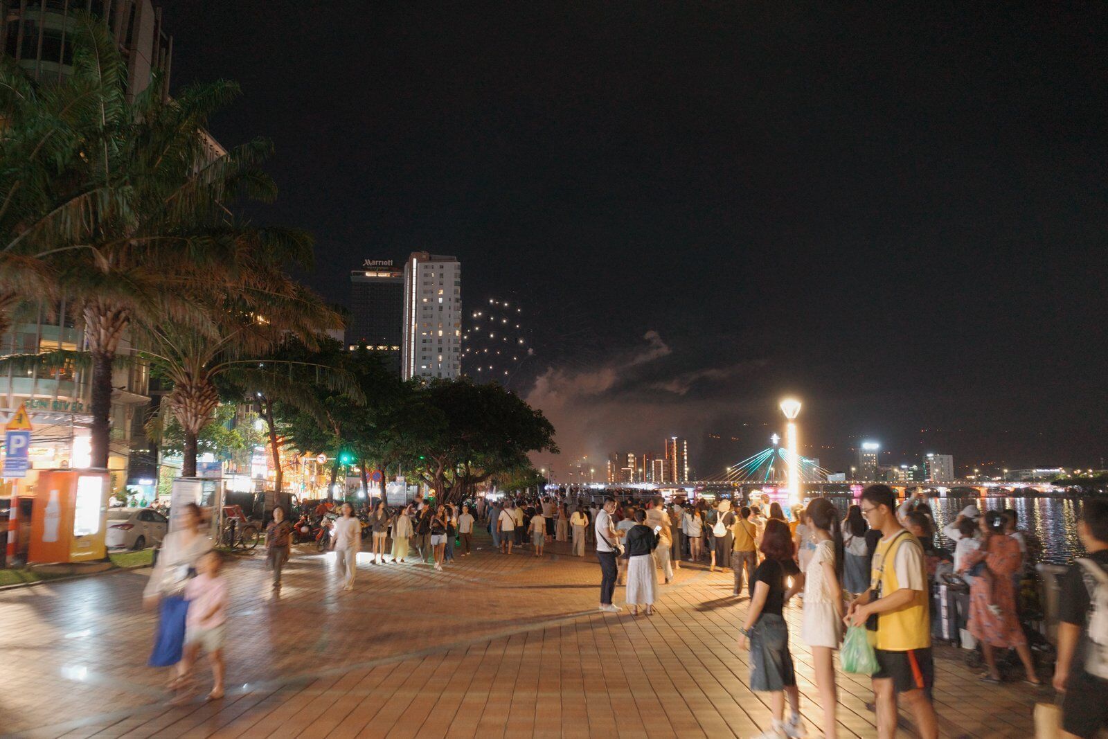 Evening crowd strolling the riverfront walkway in Da Nang under trees and city lights