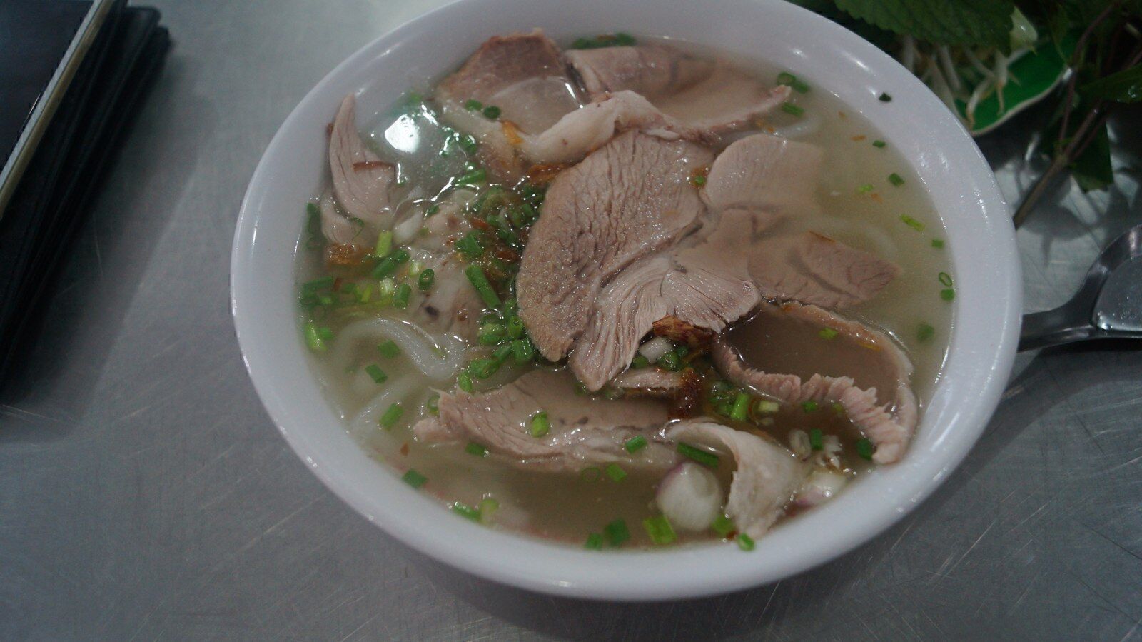 Bowl of hu tieu noodle soup with sliced pork and herbs on a metal table in Saigon