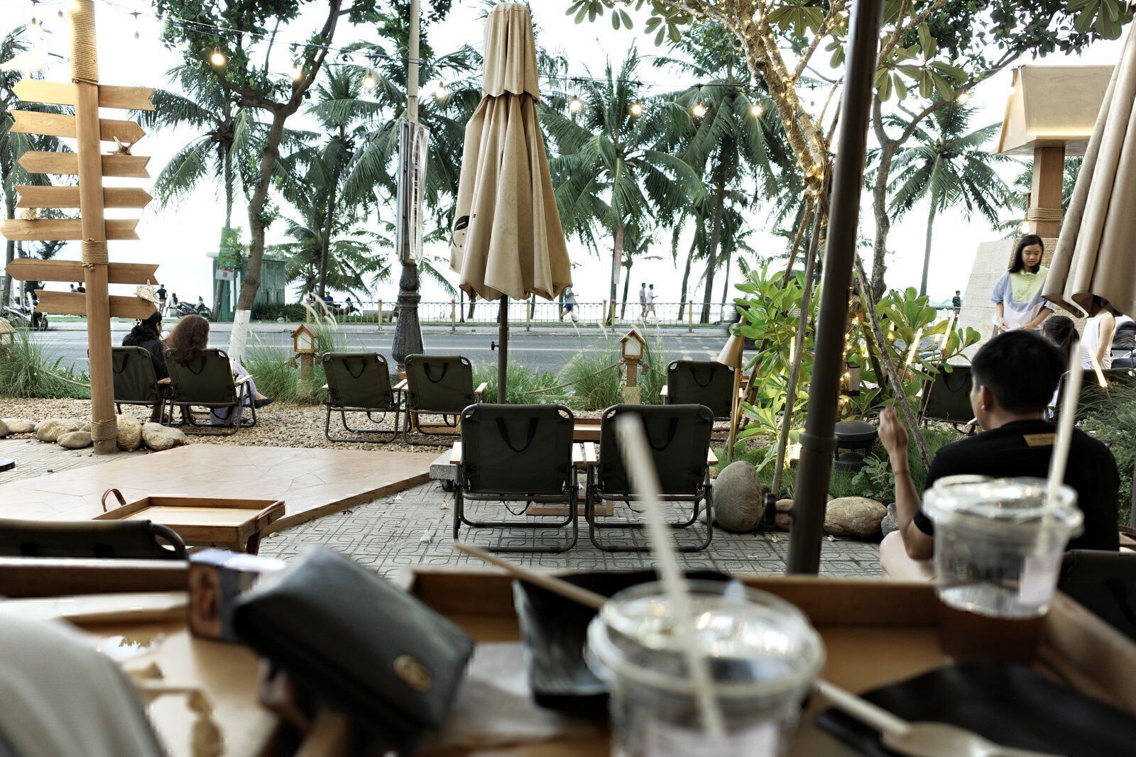 Camp chairs and iced coffees at a beachfront Vietnamese cafe with palm trees and the sea road beyond