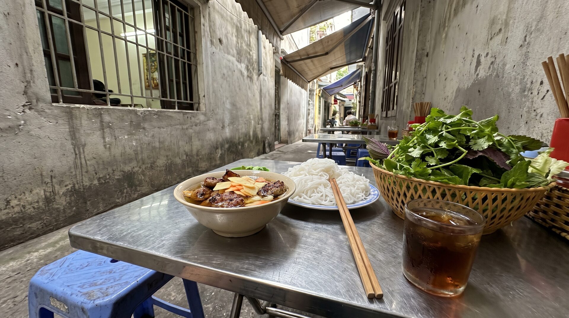 Bun cha lunch in a Hanoi alley: grilled pork in dipping broth, rice noodles, a basket of fresh herbs and iced tea on a steel table