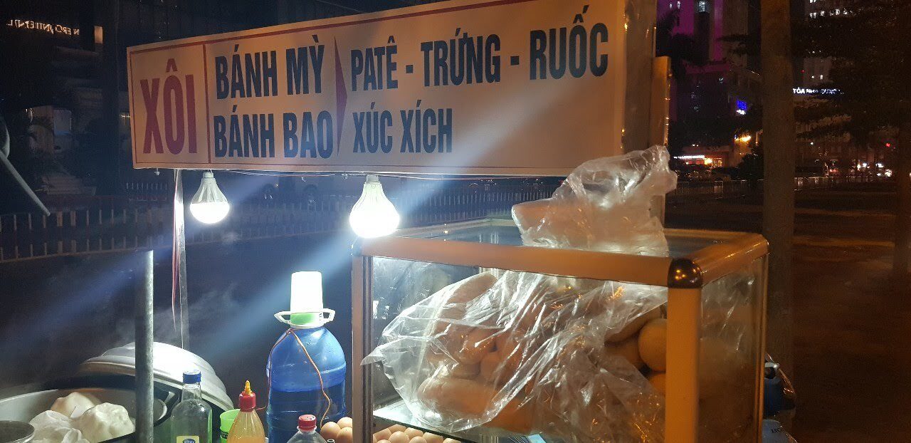 Night street cart in Hanoi selling xoi, banh my with pate, egg and ruoc under two bright bulbs