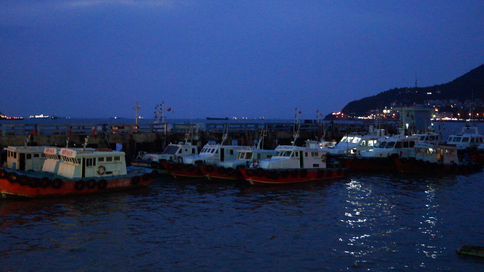 Fishing boats moored at the Vung Tau harbor pier at dusk with hill lights behind