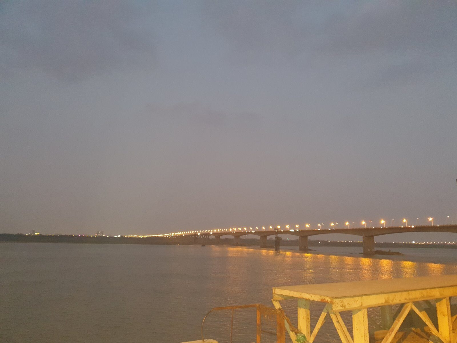 A long bridge over the Red River in Hanoi at dusk on Tet Eve, its lights just switched on, grey sky and calm water