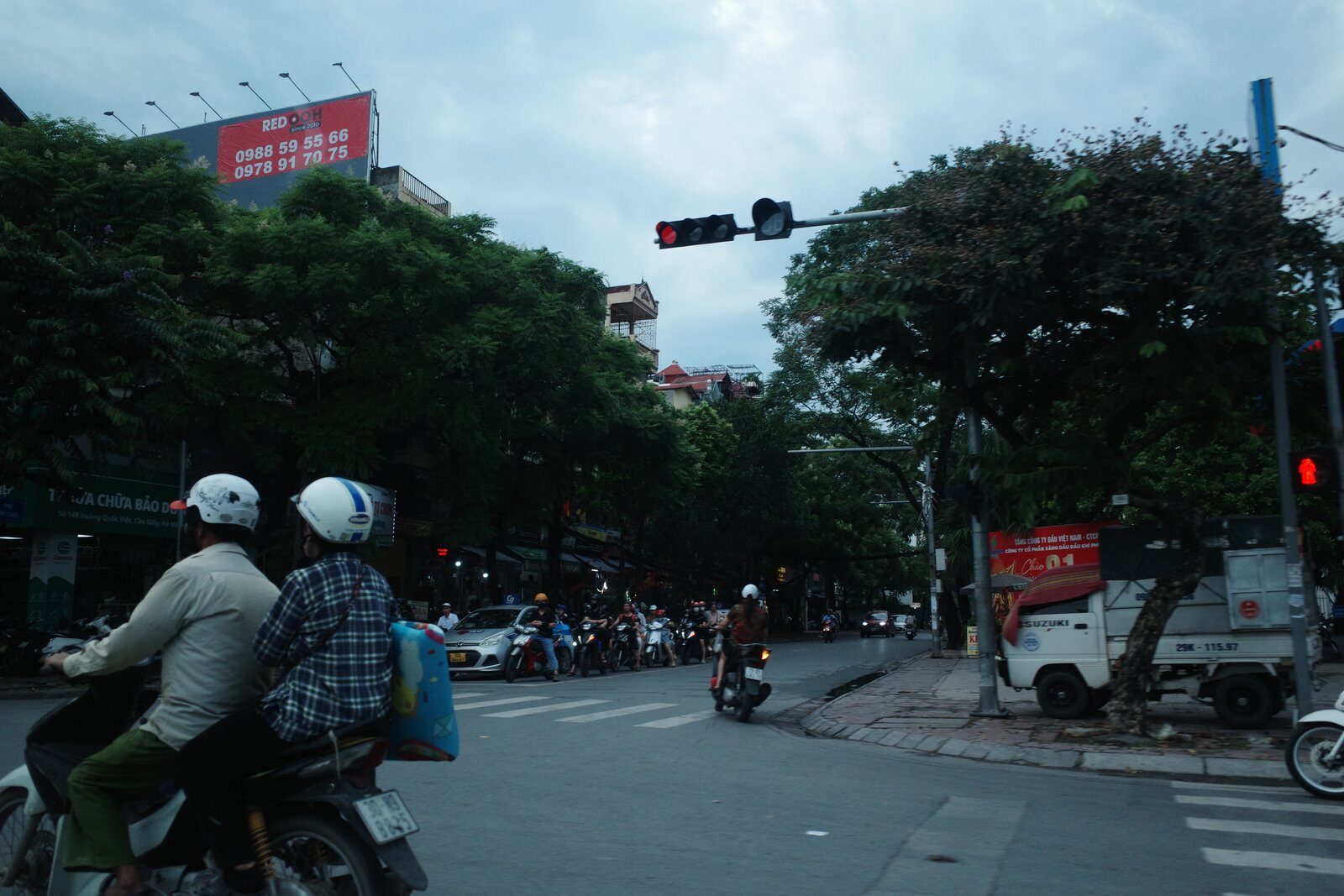 Motorbikes waiting at a traffic light on a tree-lined Hanoi boulevard in late August, grey sky and shop signs